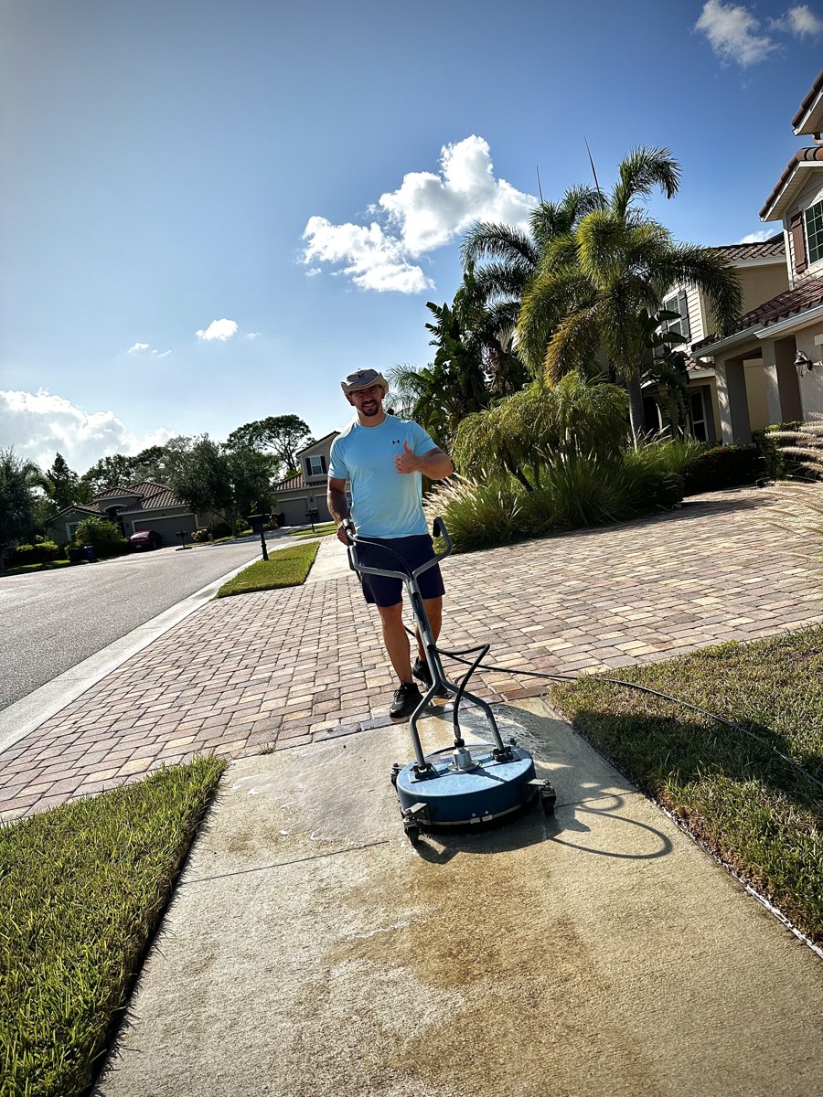 Cleansy technician giving a thumbs up next to a surface cleaner, with half the driveway already cleaned