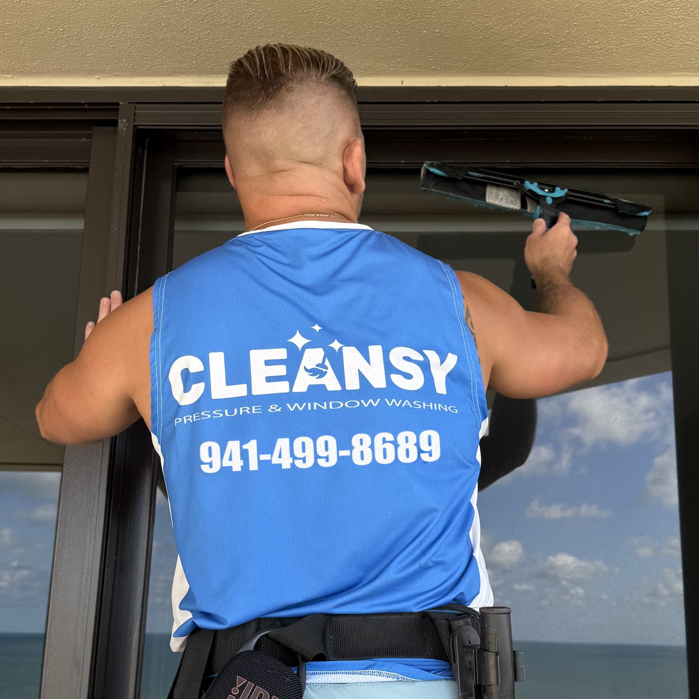 Cleansy technician in a branded blue shirt cleaning a high-rise balcony window with a squeegee, with the ocean in the background