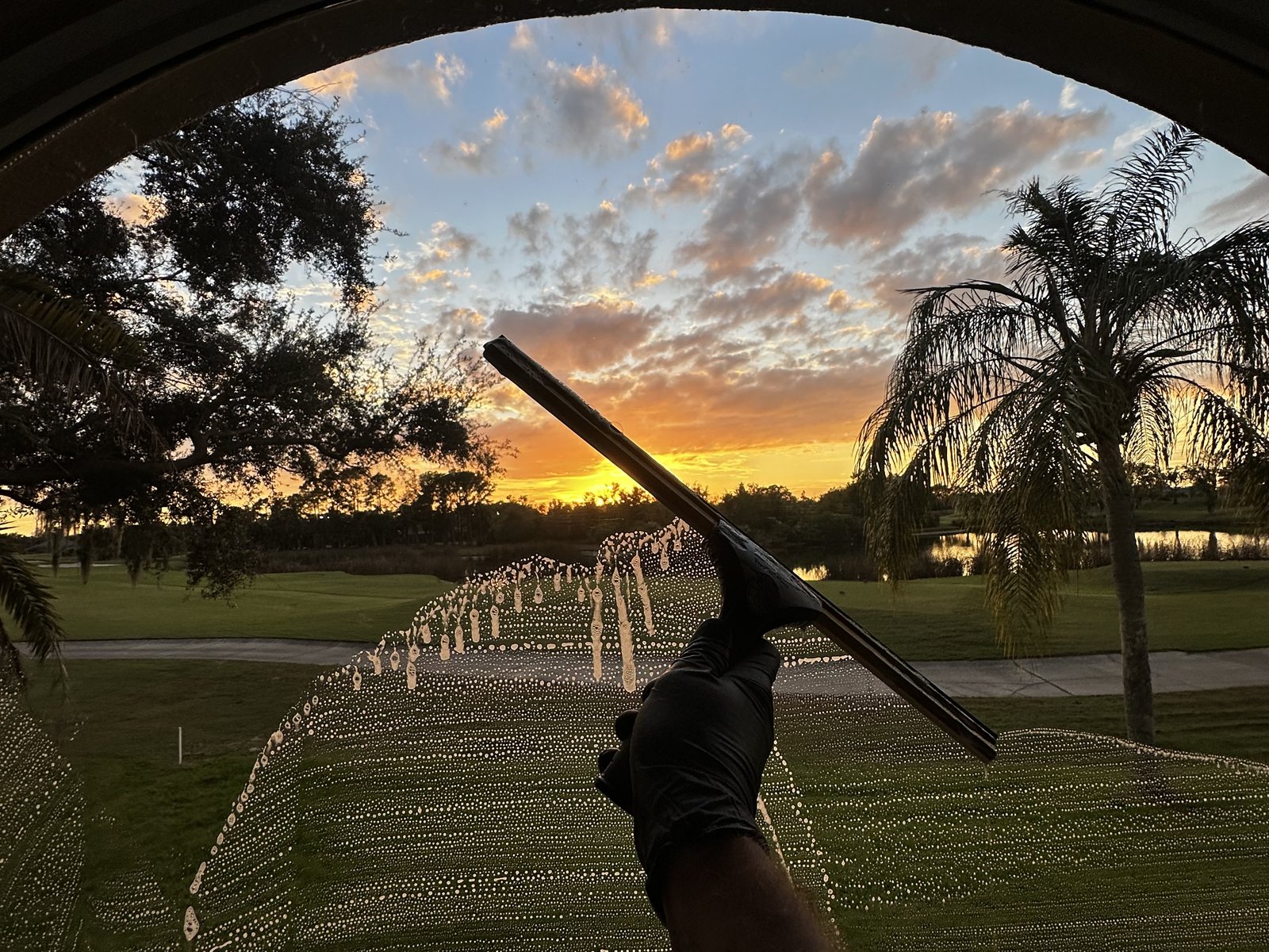 Squeegee in a gloved hand clearing an arched window with a golf course sunset behind it