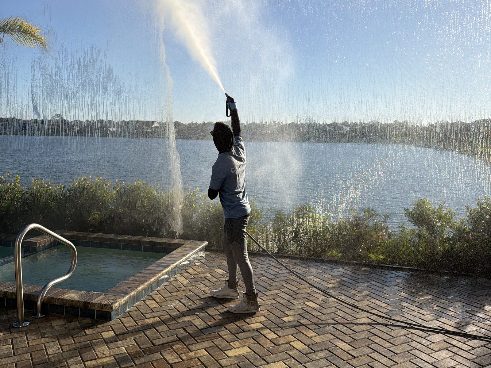 Cleansy crew member rinsing pool cage screens on a lanai beside a lake
